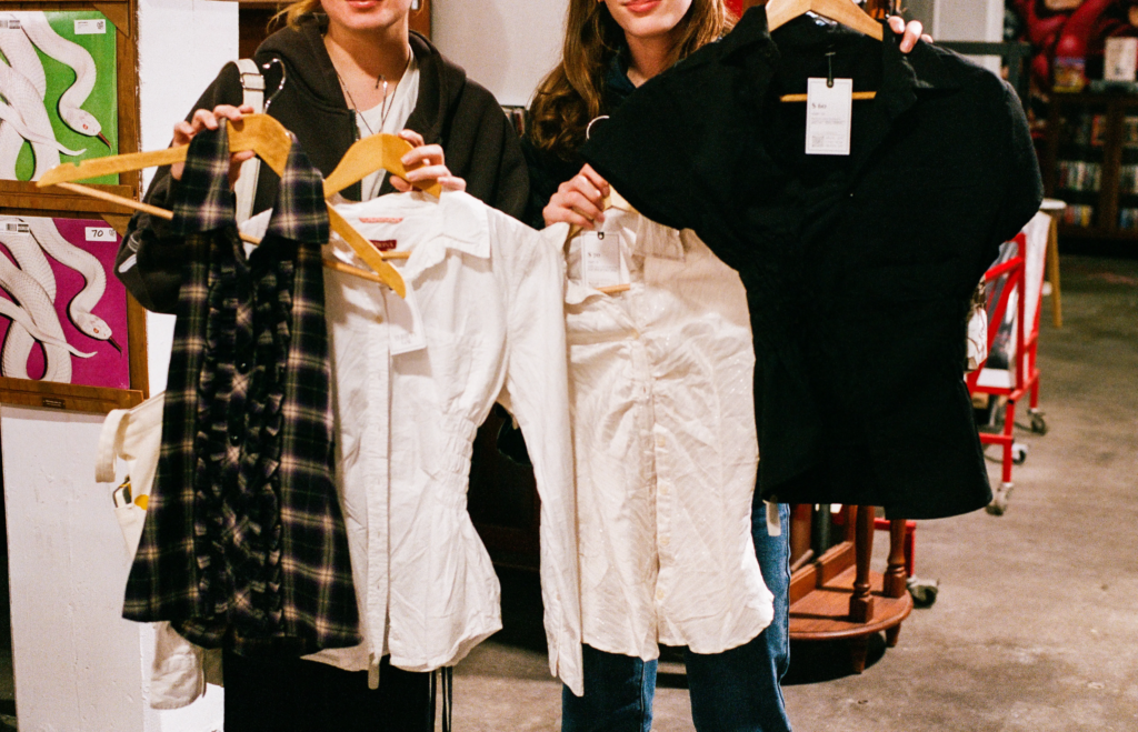 Two shoppers holding Thrifty Gal Y2K tops and preloved shirts inside My Dead Grandpa Adelaide.