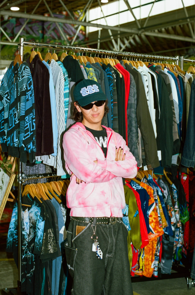 Shopper wearing a pink hoodie, baggy jeans, sunglasses and cap in front of vintage surfwear racks.
